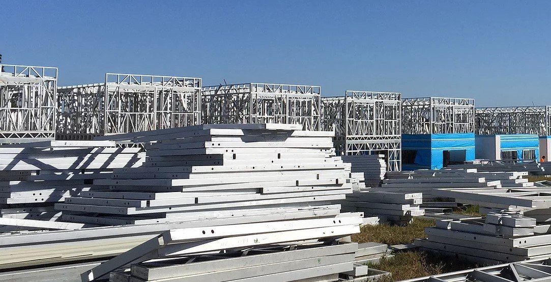 Piles of white building materials in front of a blue structure and multi-story white scaffolding under a clear sky.