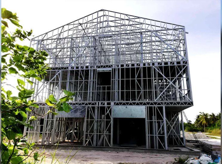 Steel frame of a two-story building under construction, exterior view with blue sky, and foliage in foreground.