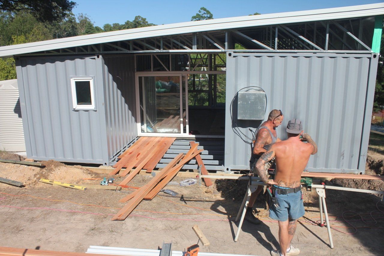 Two shirtless men building a gray shipping container home. Partially constructed, with wooden ramp and metal roof.