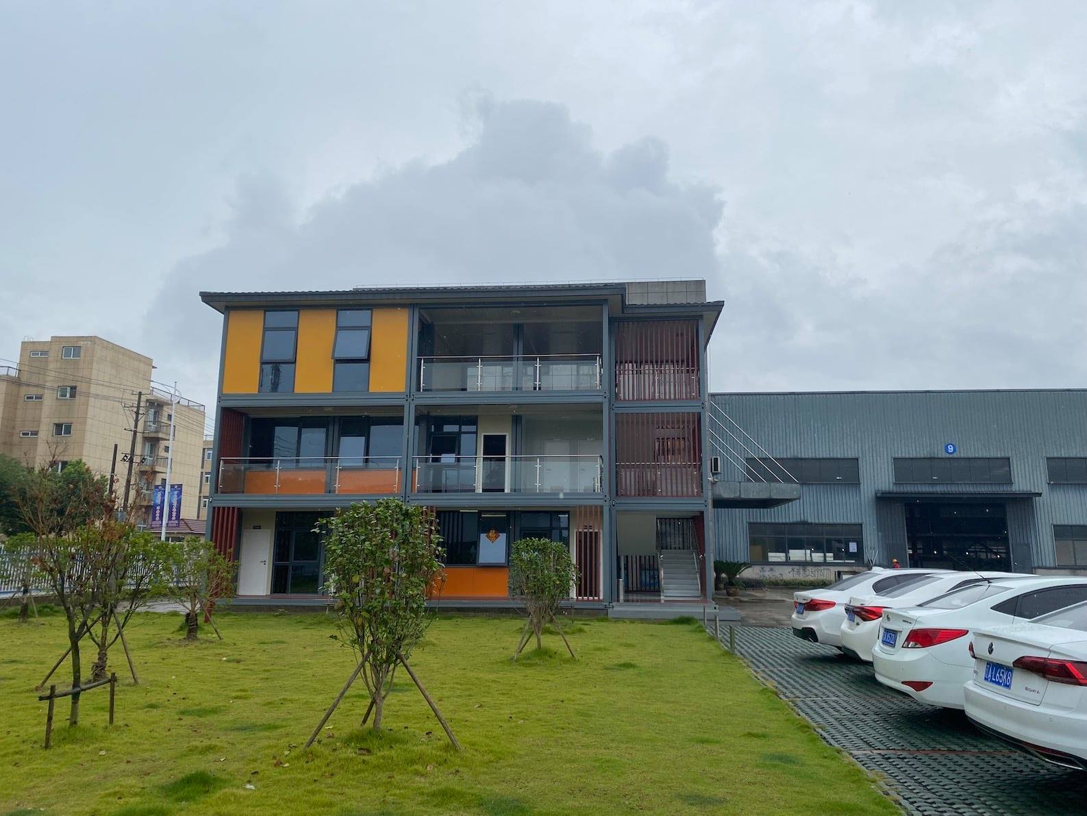 Three-story building made of shipping containers; orange and yellow accents, glass balconies, cars parked in front.
