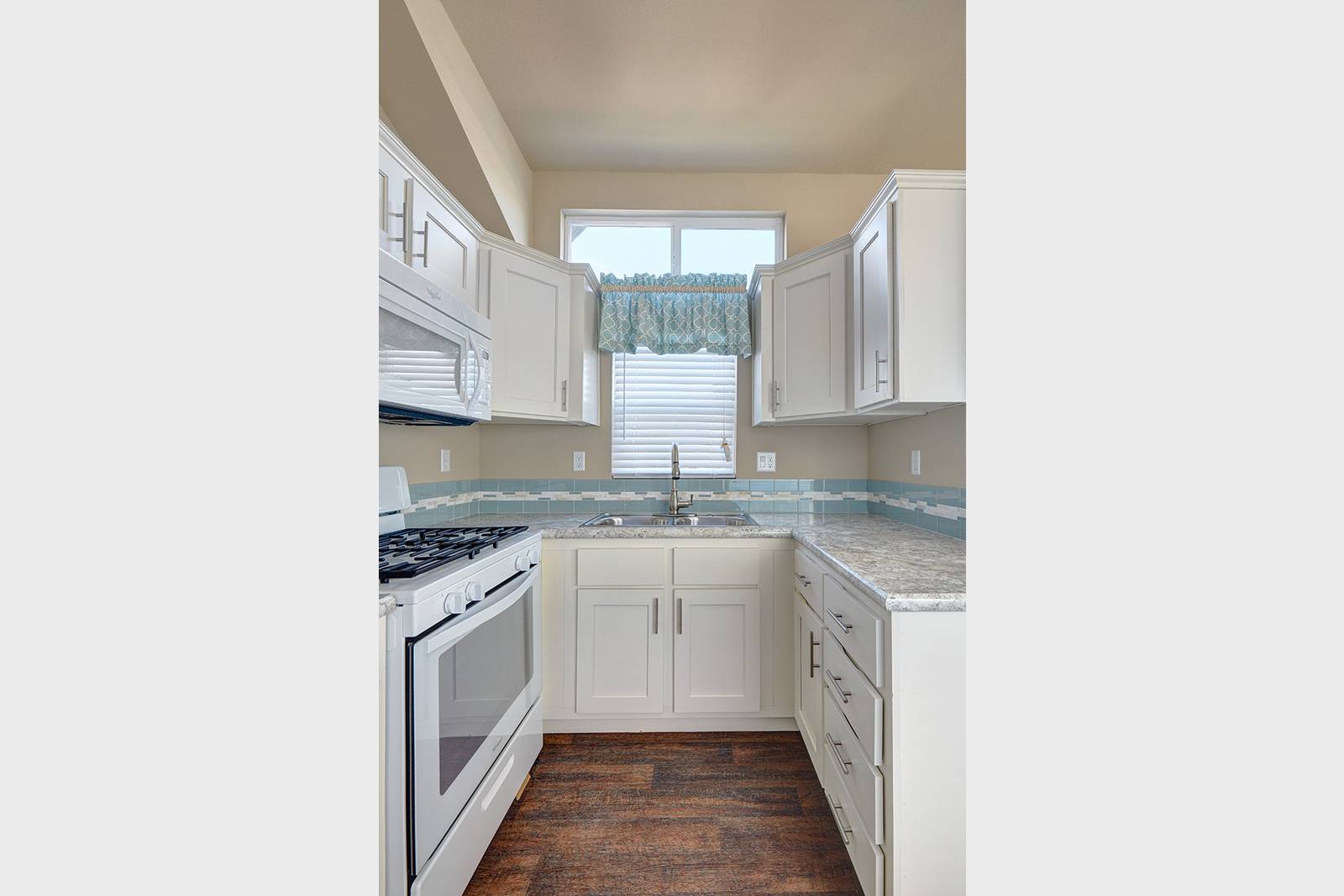 White kitchen with cabinets, stove, sink, and window with patterned valance. Brown floor.