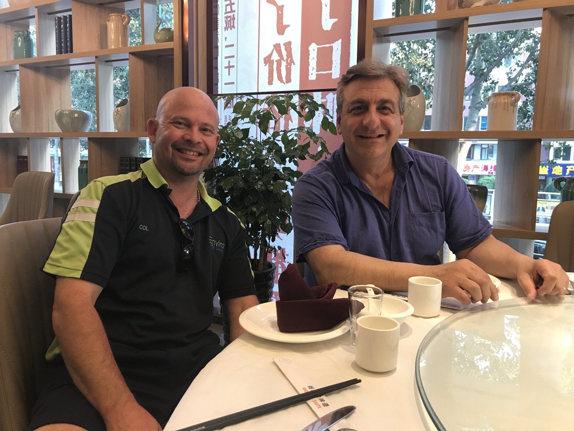 Two men smiling at a table in a restaurant. One wears a green shirt, the other a blue shirt.
