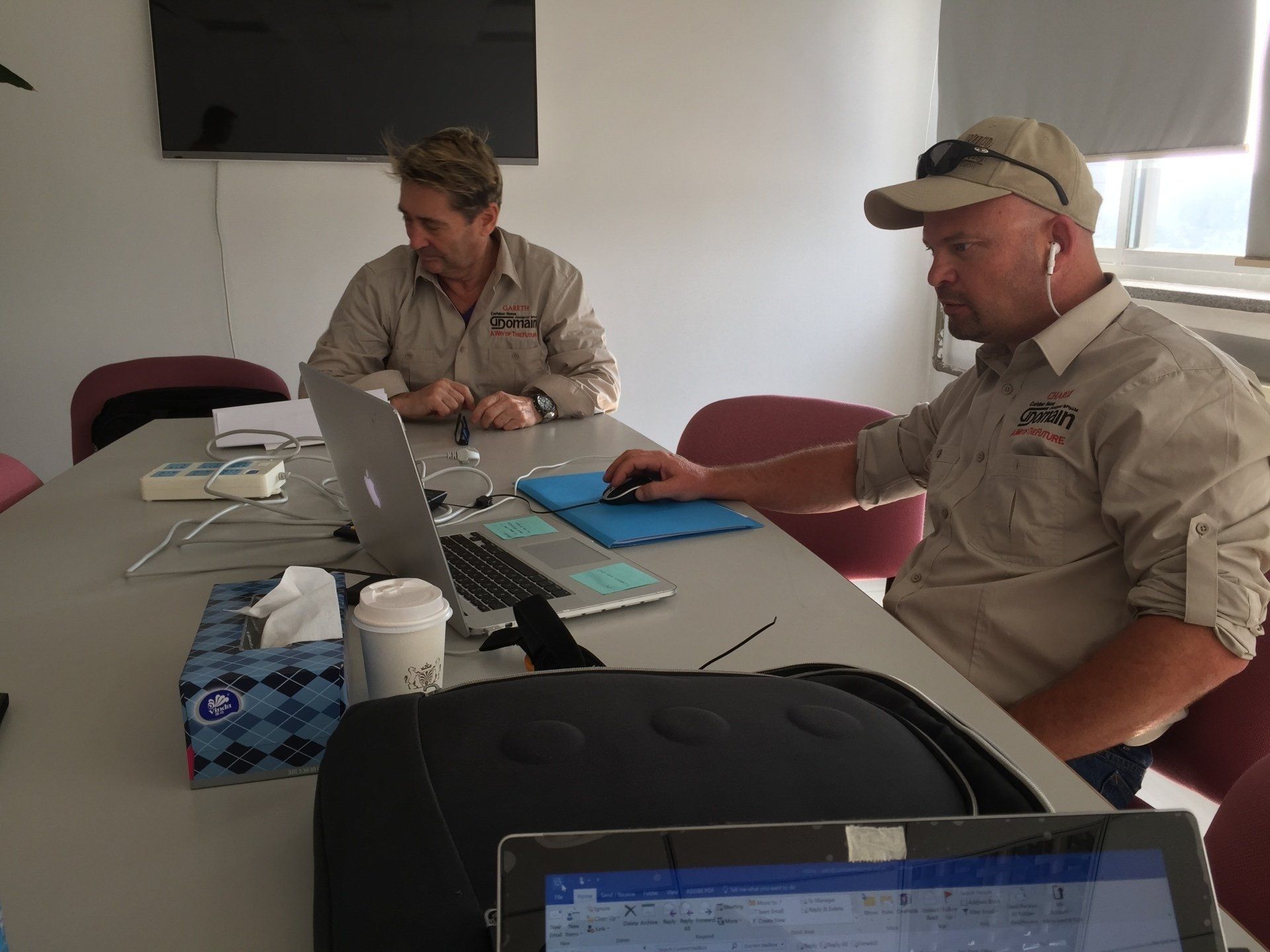 Two men in tan shirts sit at a table with a laptop, tissues, and coffee. One man uses a mouse, wearing a cap with headphones.