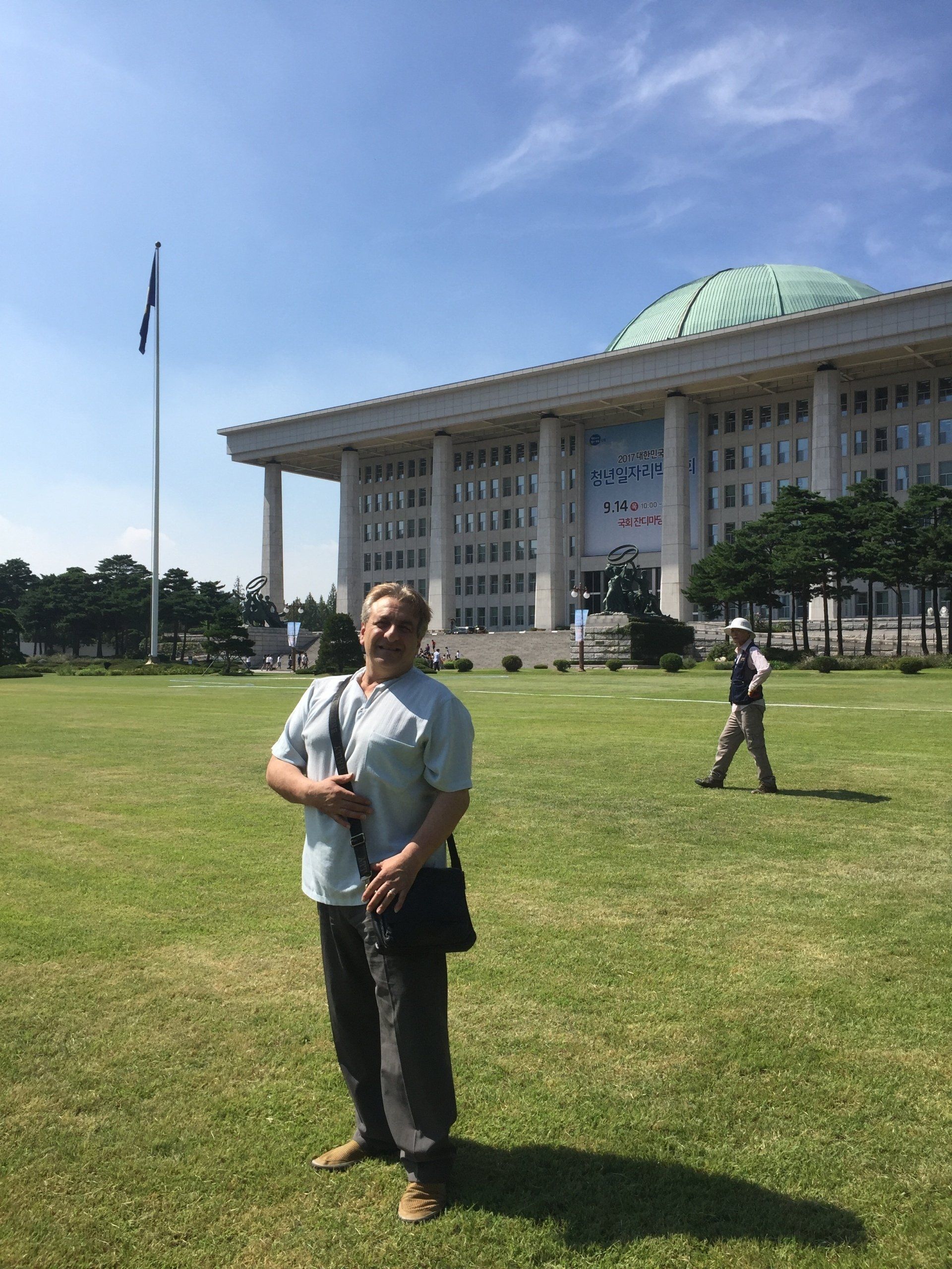 Man stands in front of a large building with a dome roof and a flag, green grass, sunny day.