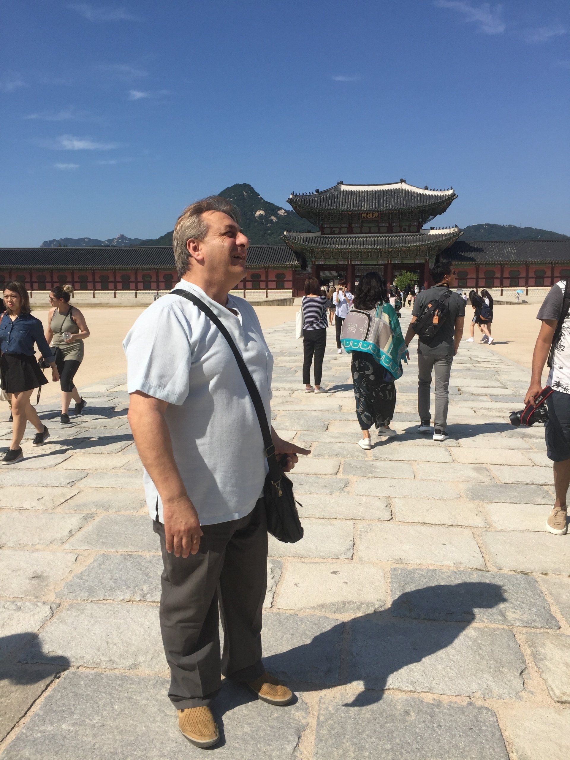 Man stands in front of a traditional building with tourists. He is wearing a light-colored shirt and looking upwards.