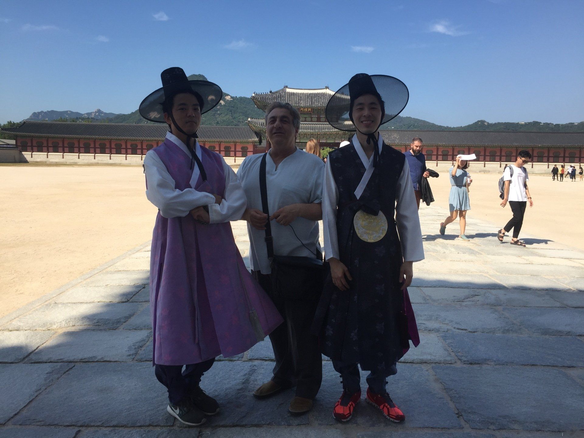 Two men in traditional Korean garb pose with a man in front of a building; outdoor setting.