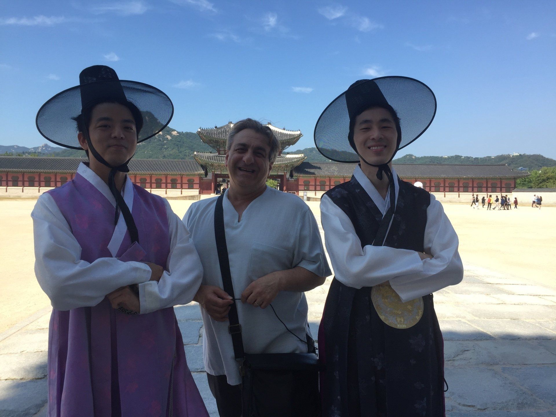 Three people pose in front of a Korean palace. Two wear traditional hats and robes, one in white shirt.