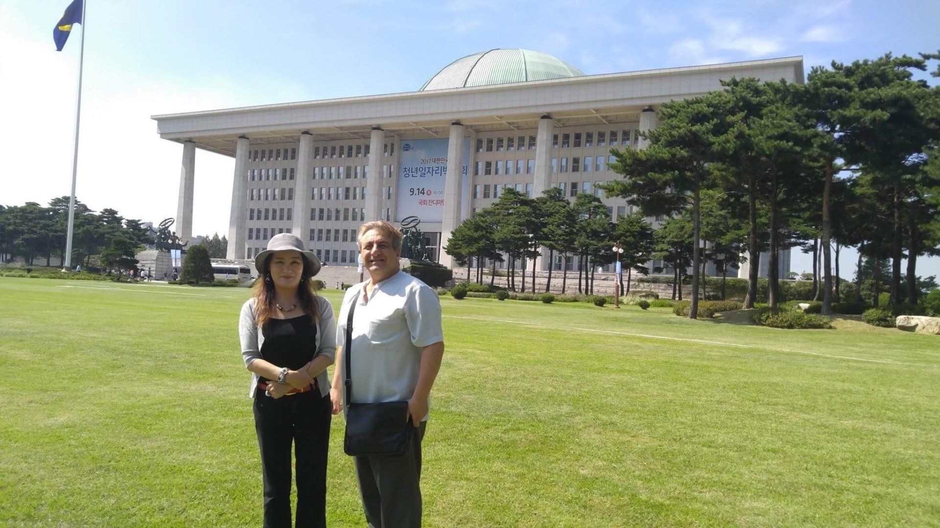 Two people stand in front of a large building with a dome roof and columns. Green lawn. Sunny day.