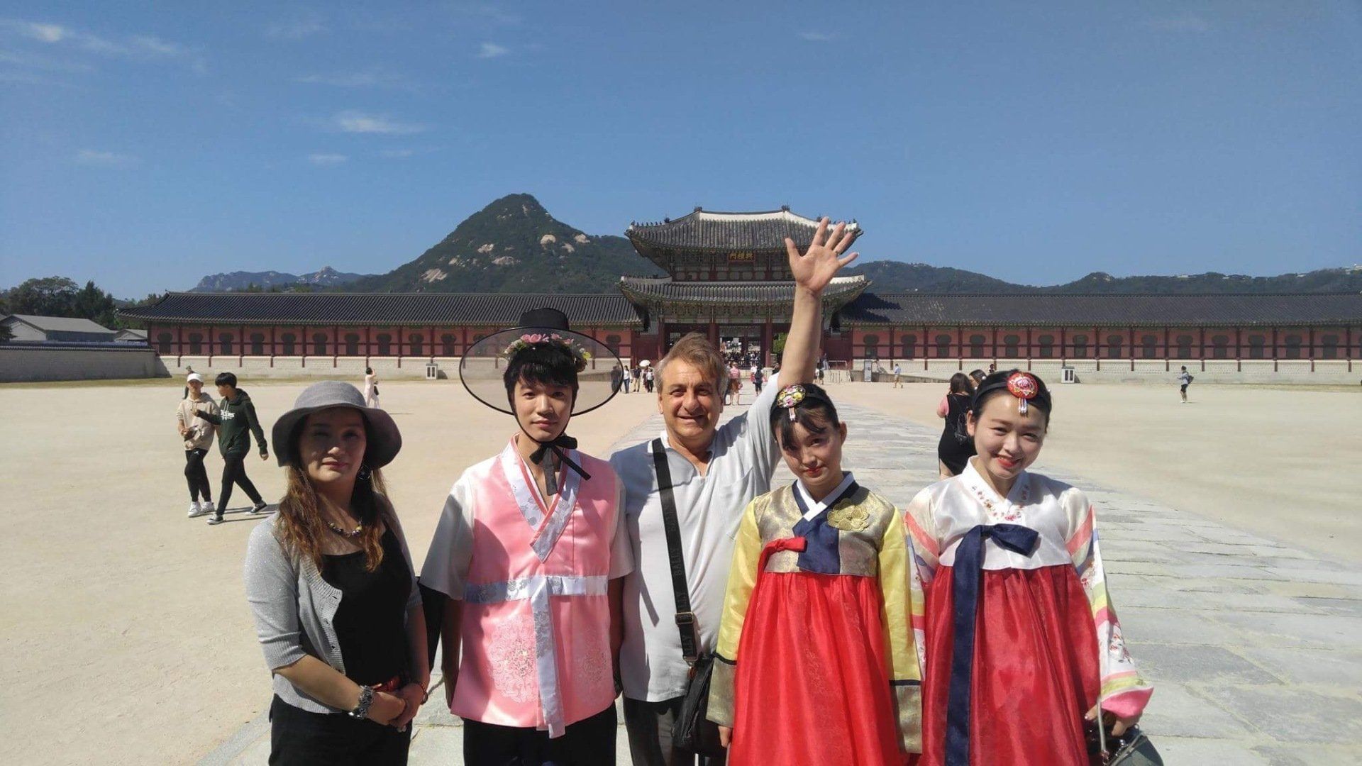 Group of five people at Gyeongbokgung Palace, Seoul. Person in the center waves, others wear Korean clothing. Sunny day.