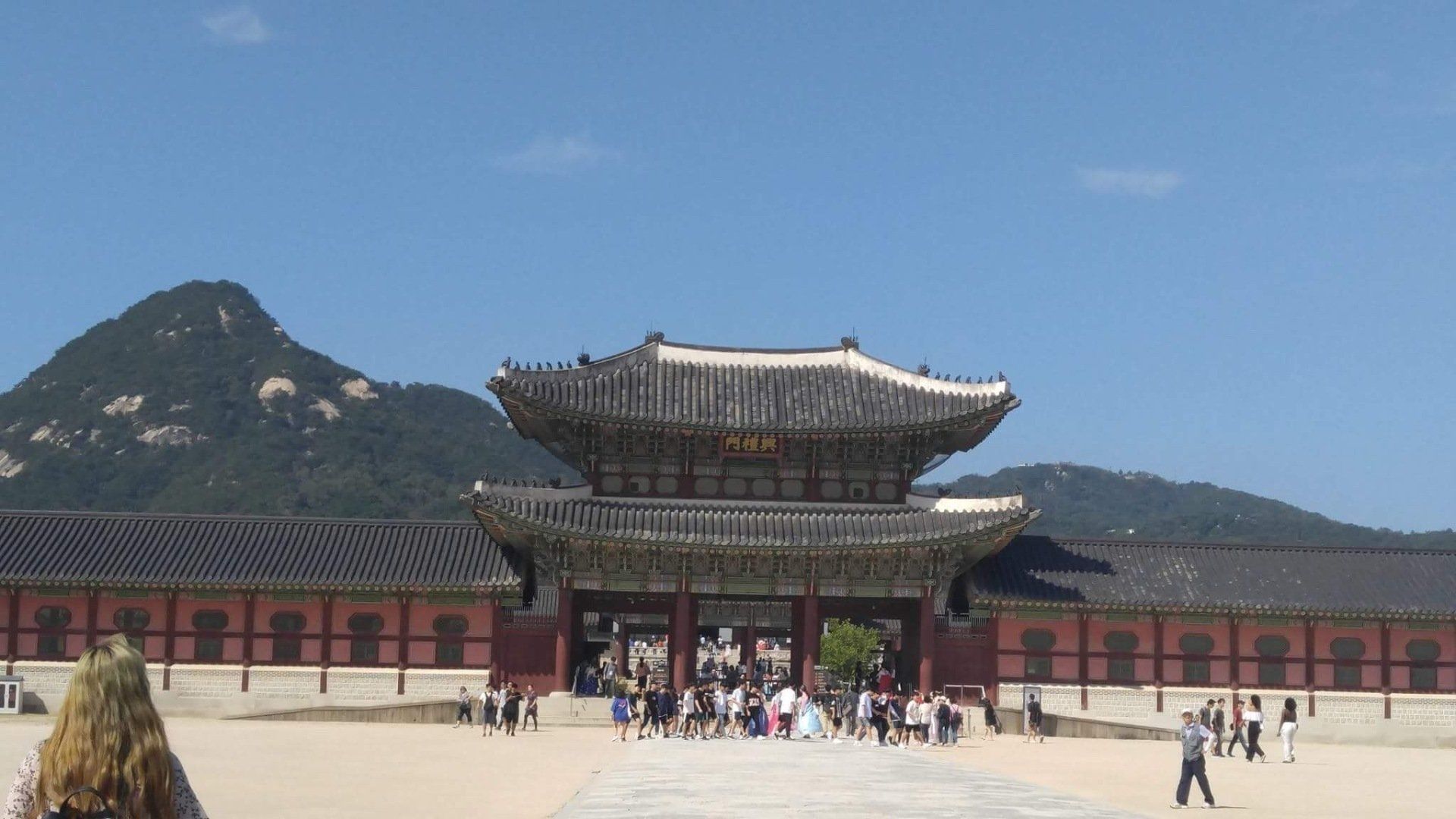 Traditional Korean palace gate with visitors under a blue sky, mountains in background.