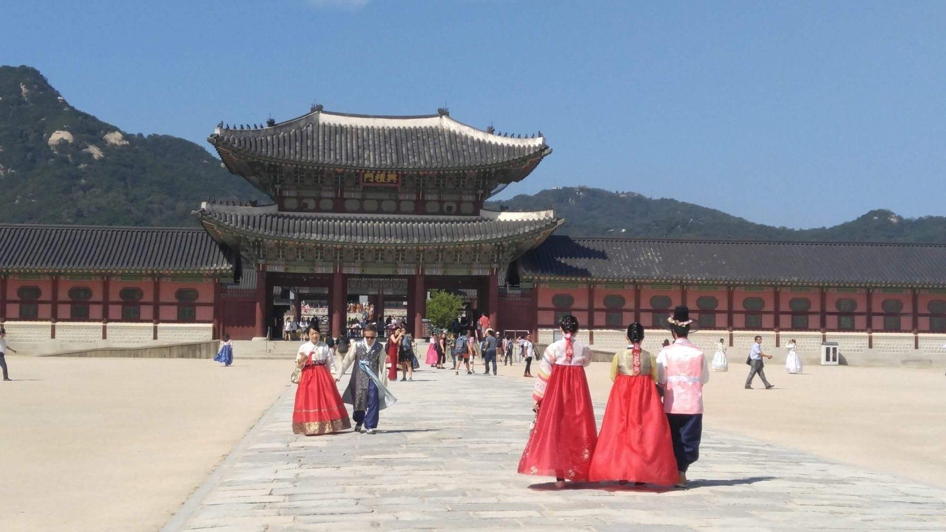 People in traditional Korean hanbok attire walk towards a palace gate; blue sky, mountains.