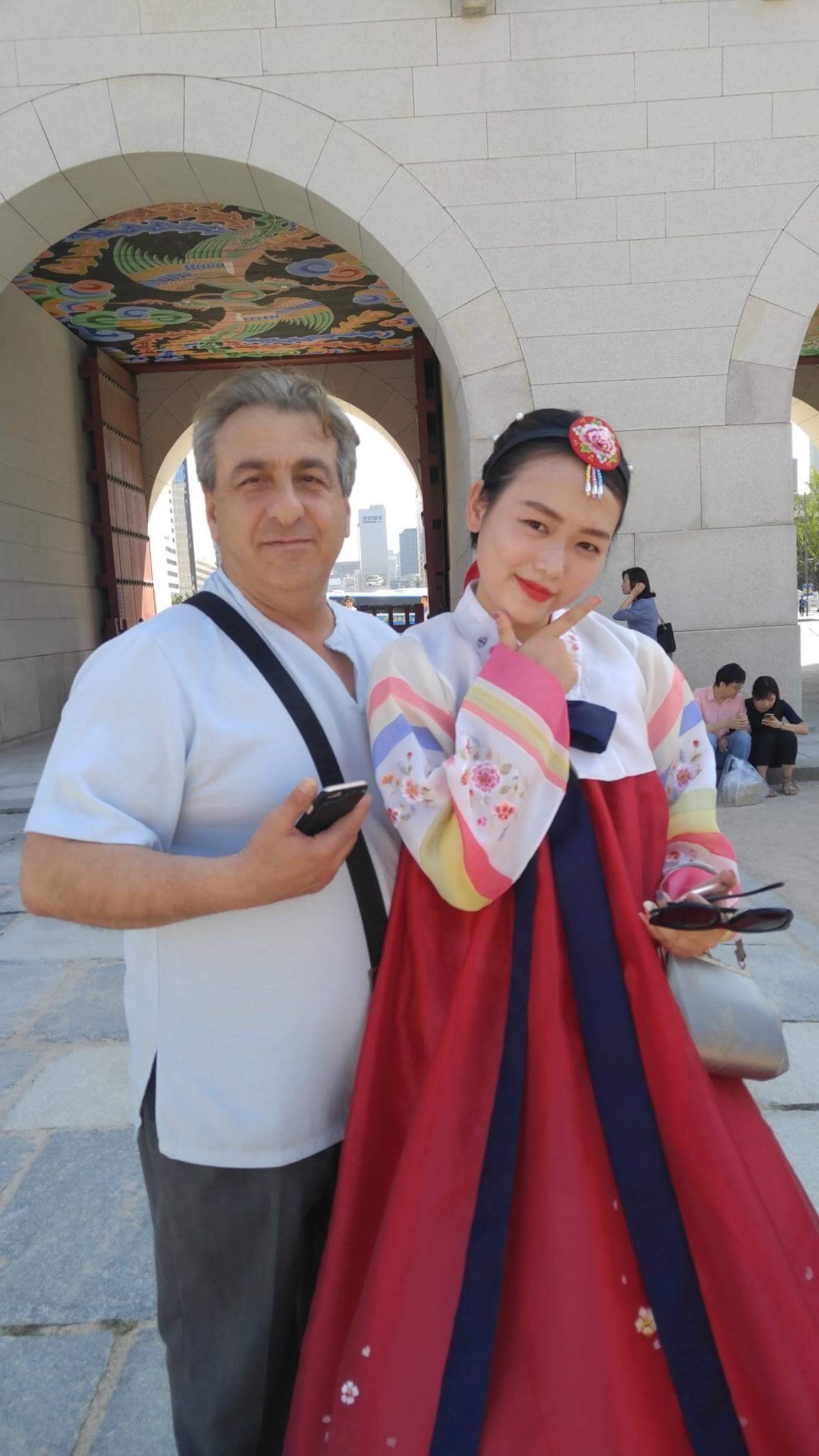 Man and woman in Korean hanbok pose in front of a stone archway. Woman smiles and gestures.