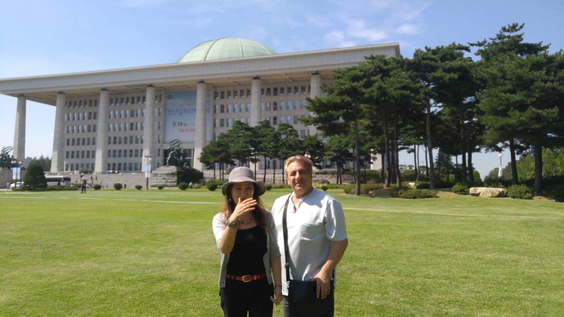 Two people stand on grass in front of a large building with a green dome; bright day.