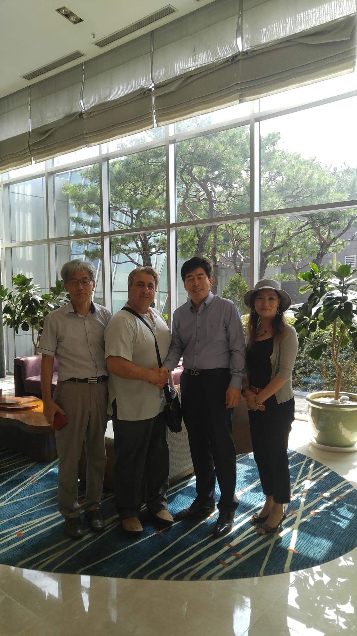 Four people pose indoors in front of a window. Two men shake hands. Trees visible outside.