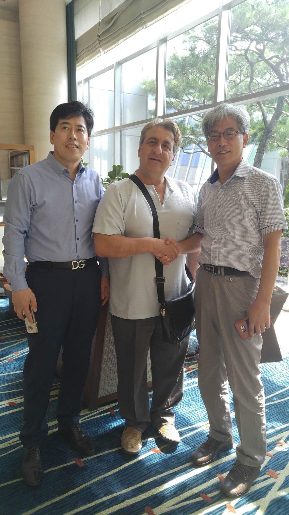 Three men shaking hands indoors near windows. One wears light shirt, another a dark button-down, and the third a patterned short-sleeved shirt.