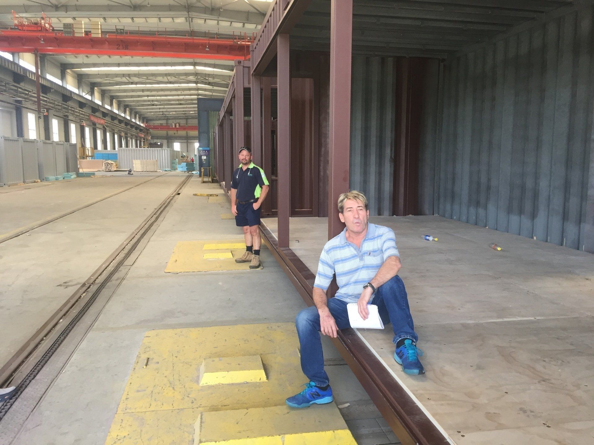 Two men inside large warehouse, near a steel frame. One seated, one standing, looking toward camera.
