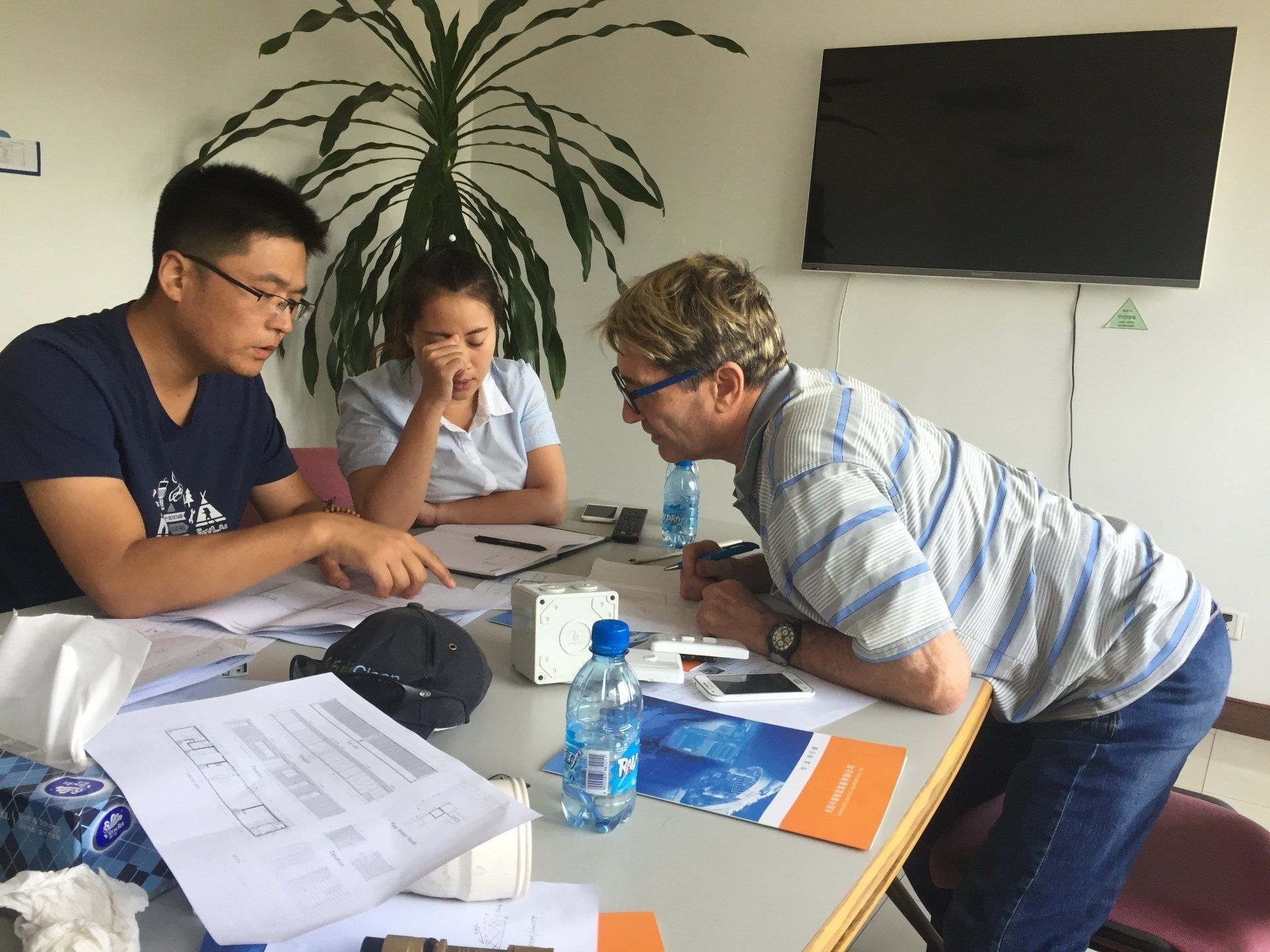 Three people in a meeting, papers and a small device on a table. One man points, others observe.
