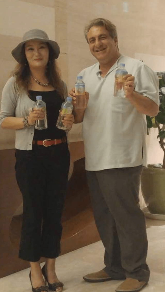Woman and man holding bottles, standing indoors. The woman wears a hat, the man smiles.