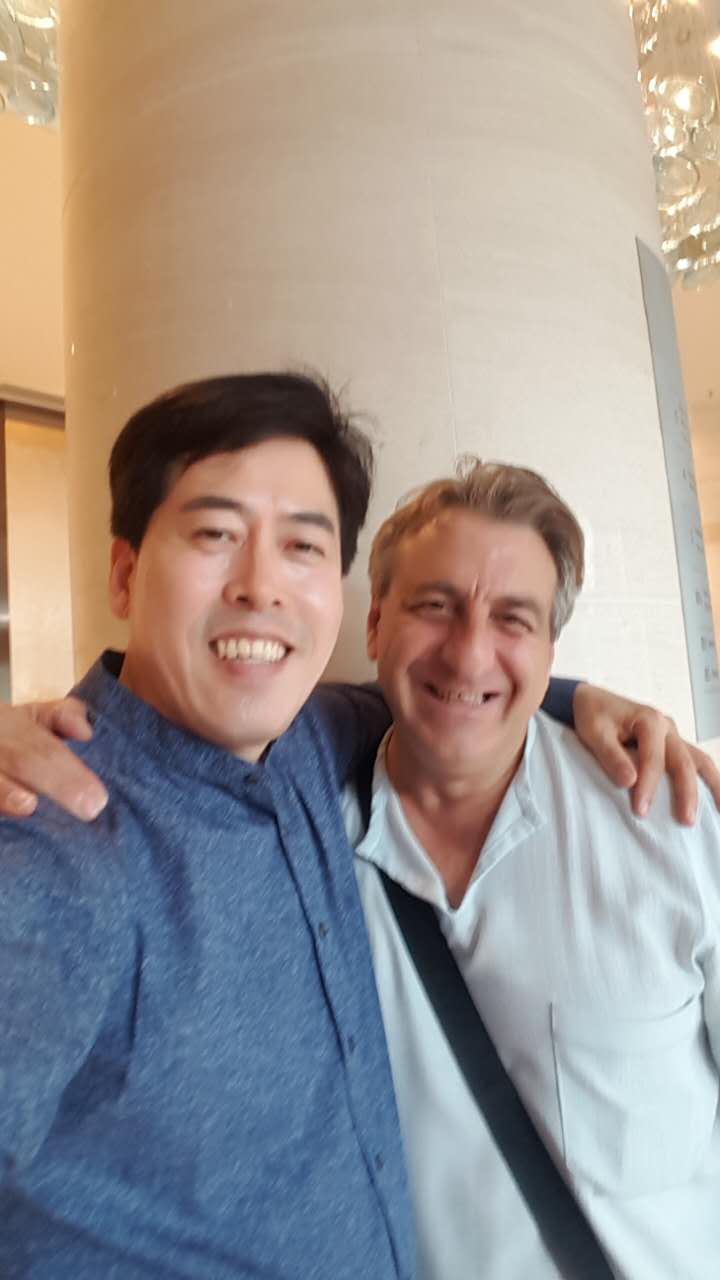 Two men smiling, arm around each other, pose for a selfie in front of a modern building column.