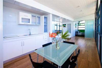Modern white kitchen with glass dining table, wooden floor, and a view of another room.