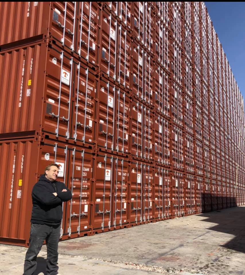 Man standing in front of tall wall of stacked, rust-colored shipping containers; sunny outdoor setting.