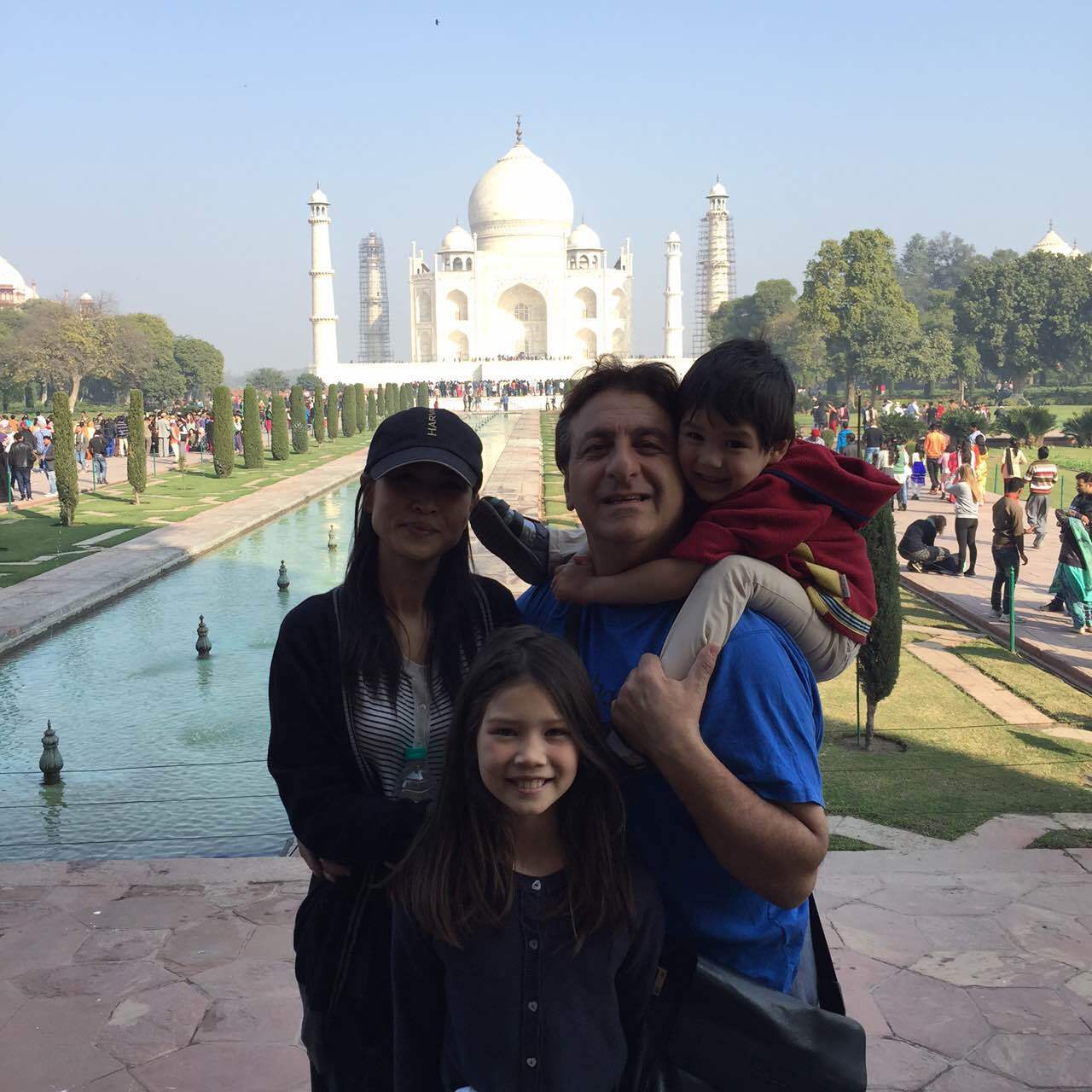 Family poses in front of the Taj Mahal. Man holds child on shoulders; woman and daughter stand near.