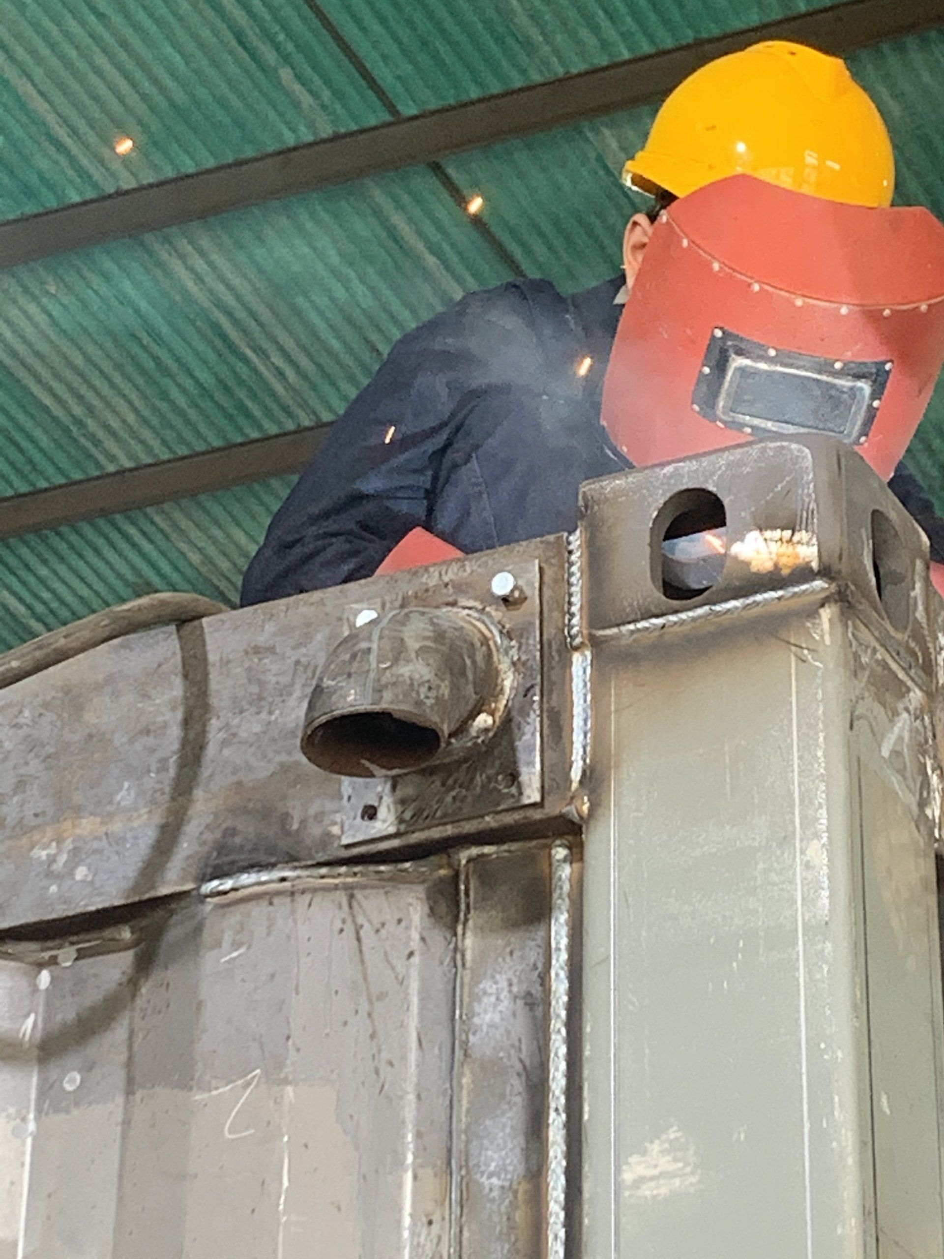Welder in a yellow hard hat and red mask, welding metal outdoors.