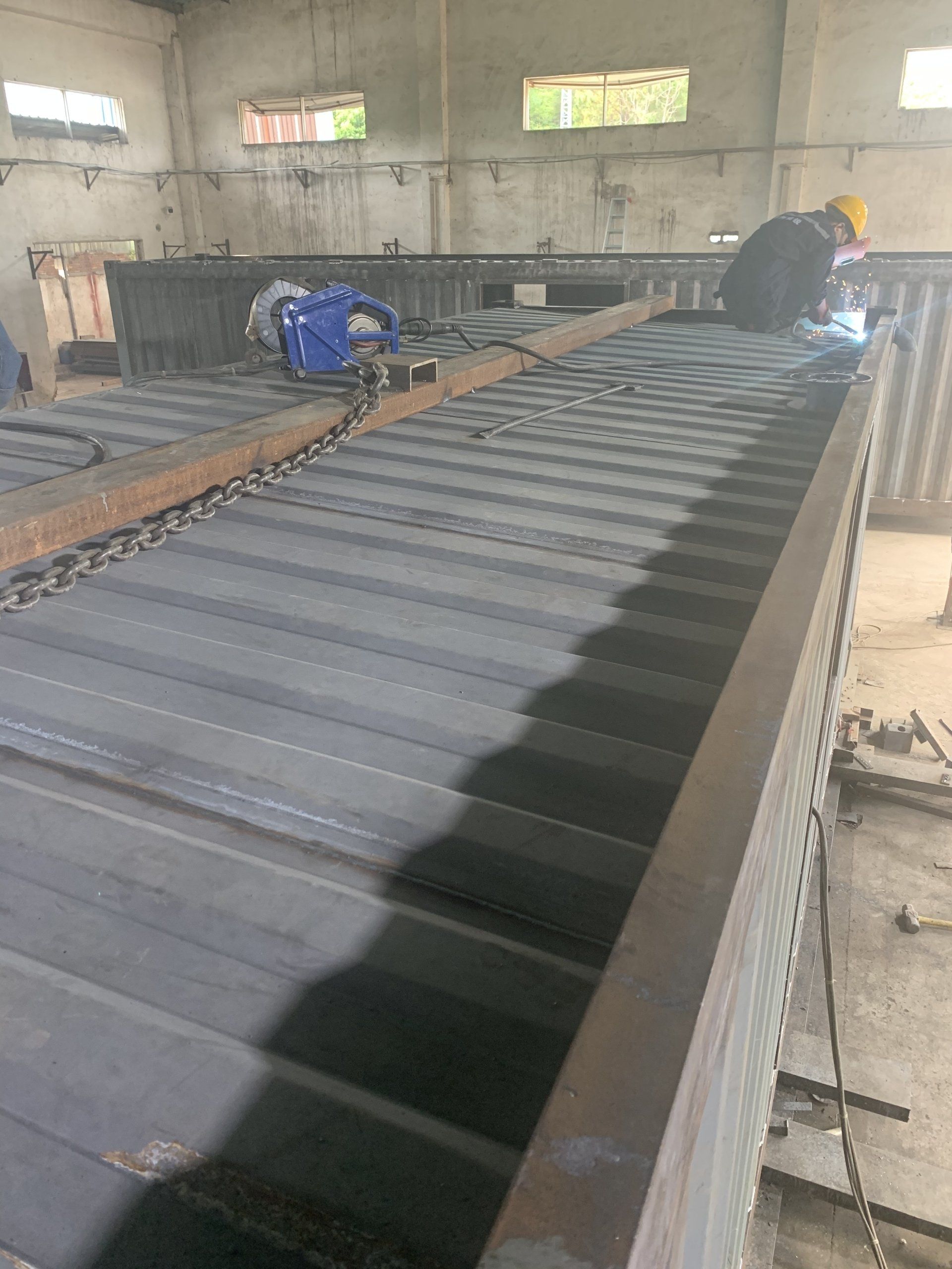 Worker welding metal structure in a factory; long, dark metal beams.