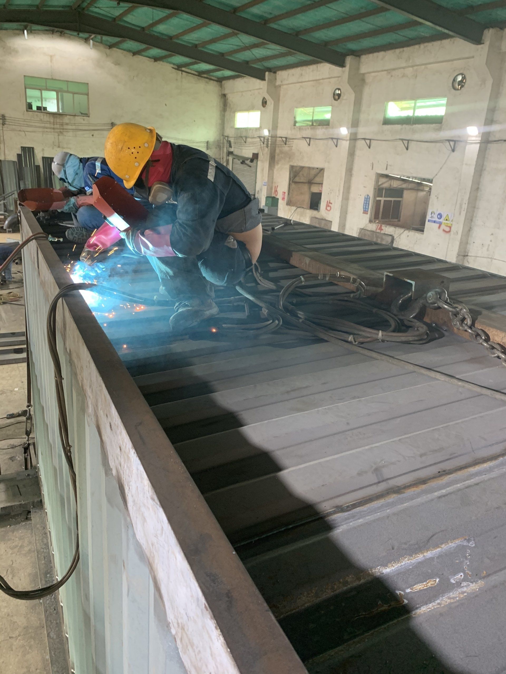 Two welders in protective gear welding metal structures in a large industrial setting.