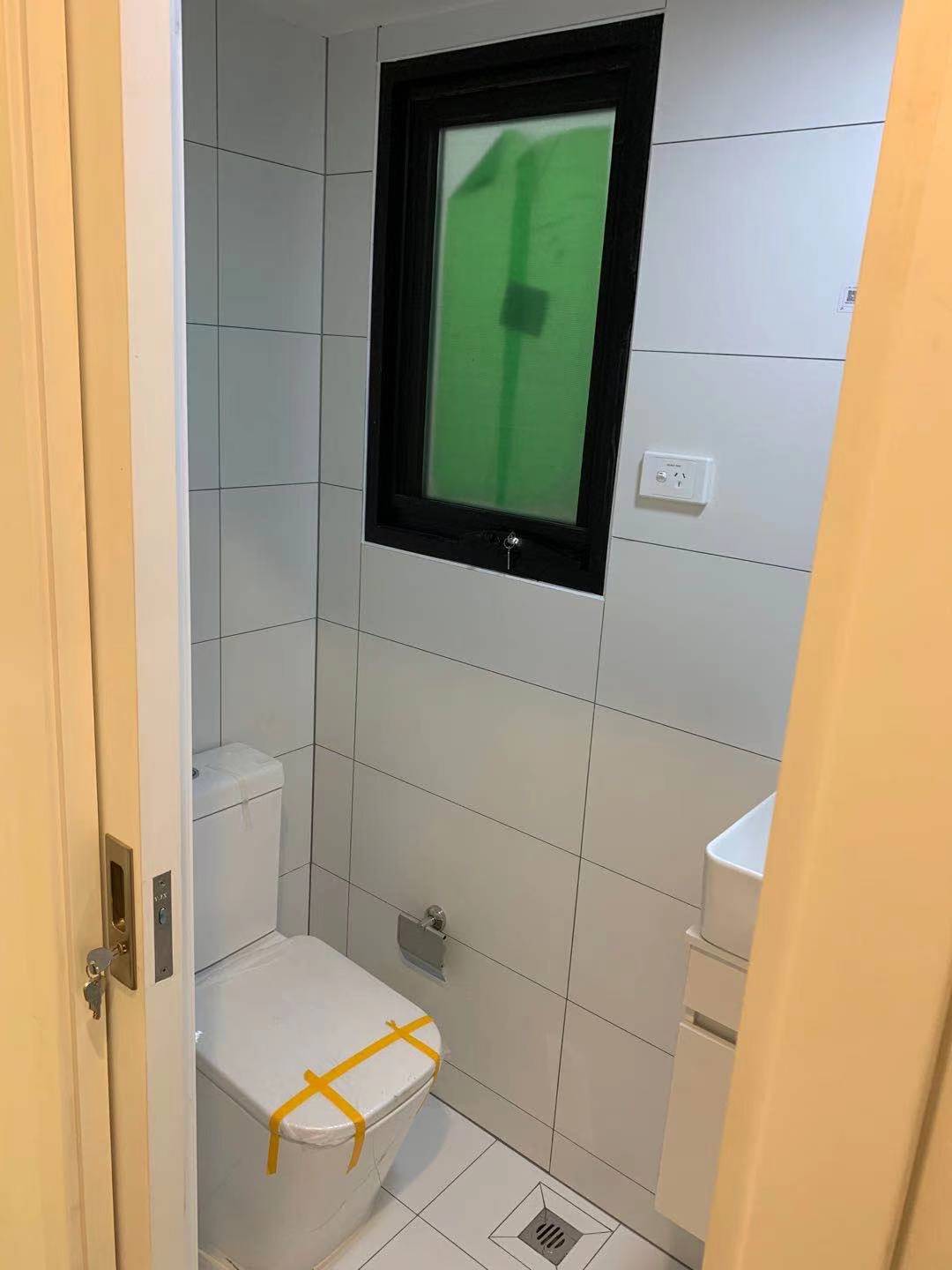 Small bathroom with white tiled walls, toilet, window with green tint, and a small sink.