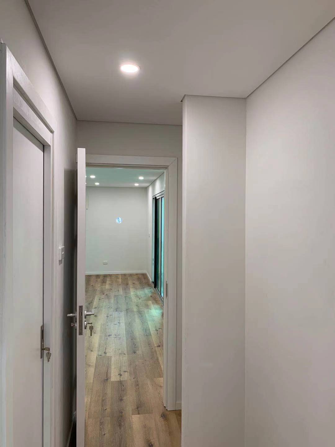 Hallway with white walls, wooden floor, and open doorway leading to a room.