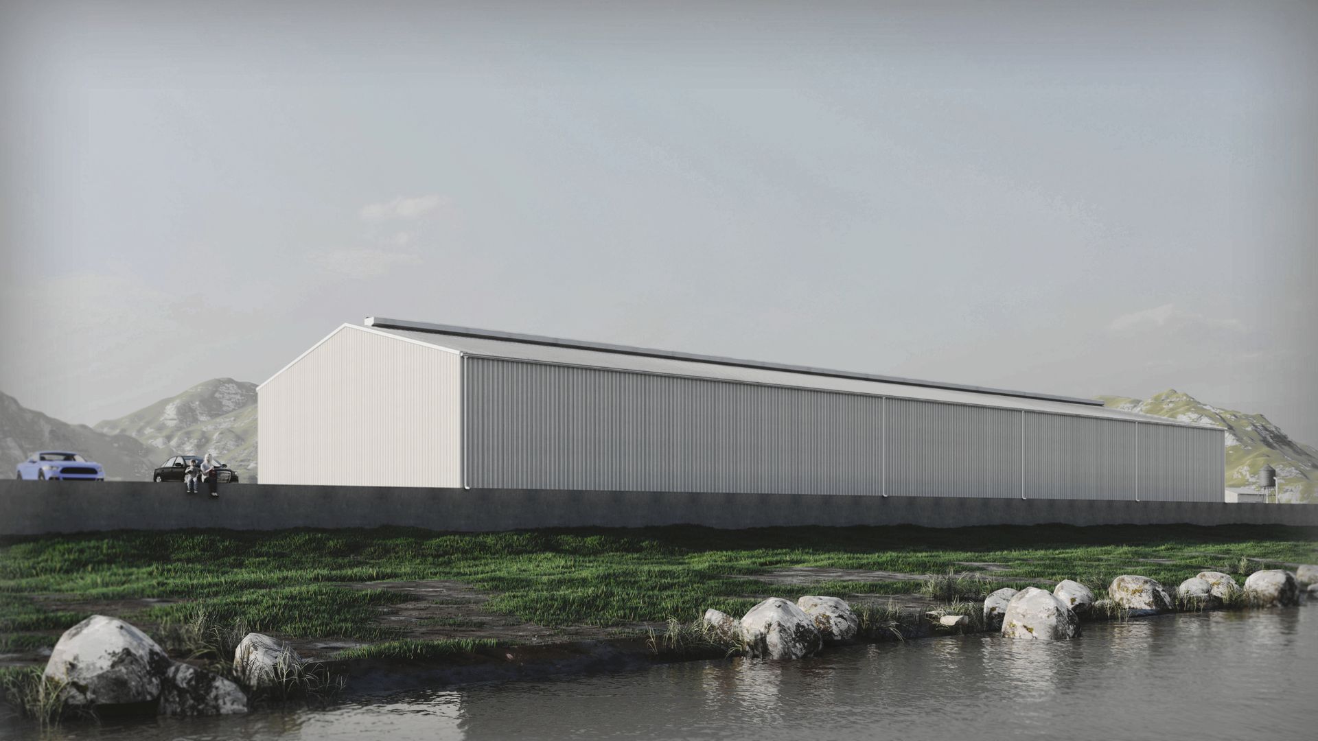 Long, white industrial building along a river, with mountains in the background.