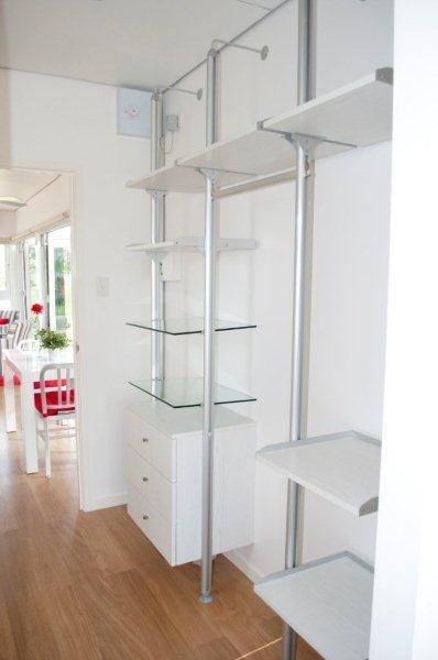 White and silver adjustable shelving unit on a white wall, with a white dresser and glass shelves.