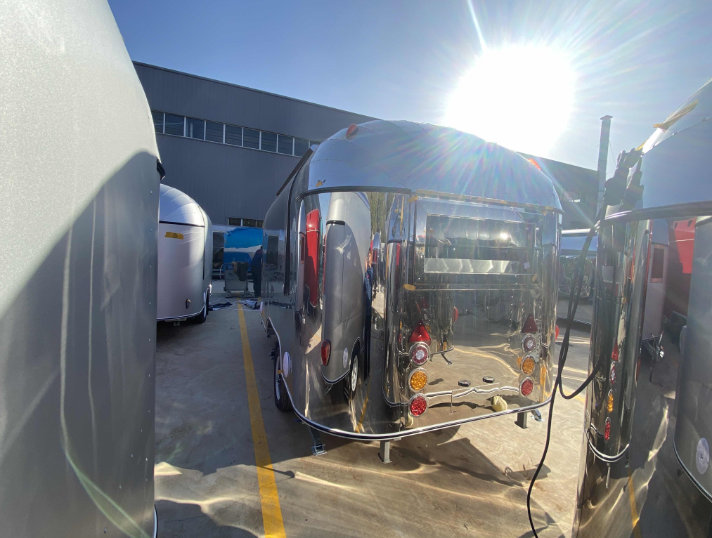 Shiny silver Airstream trailer parked outdoors in sunlight, next to other trailers.