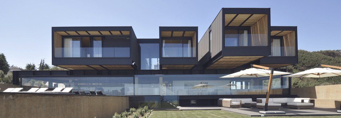 Modern black house with glass and wood design, cantilevered over a green lawn with a swimming pool.