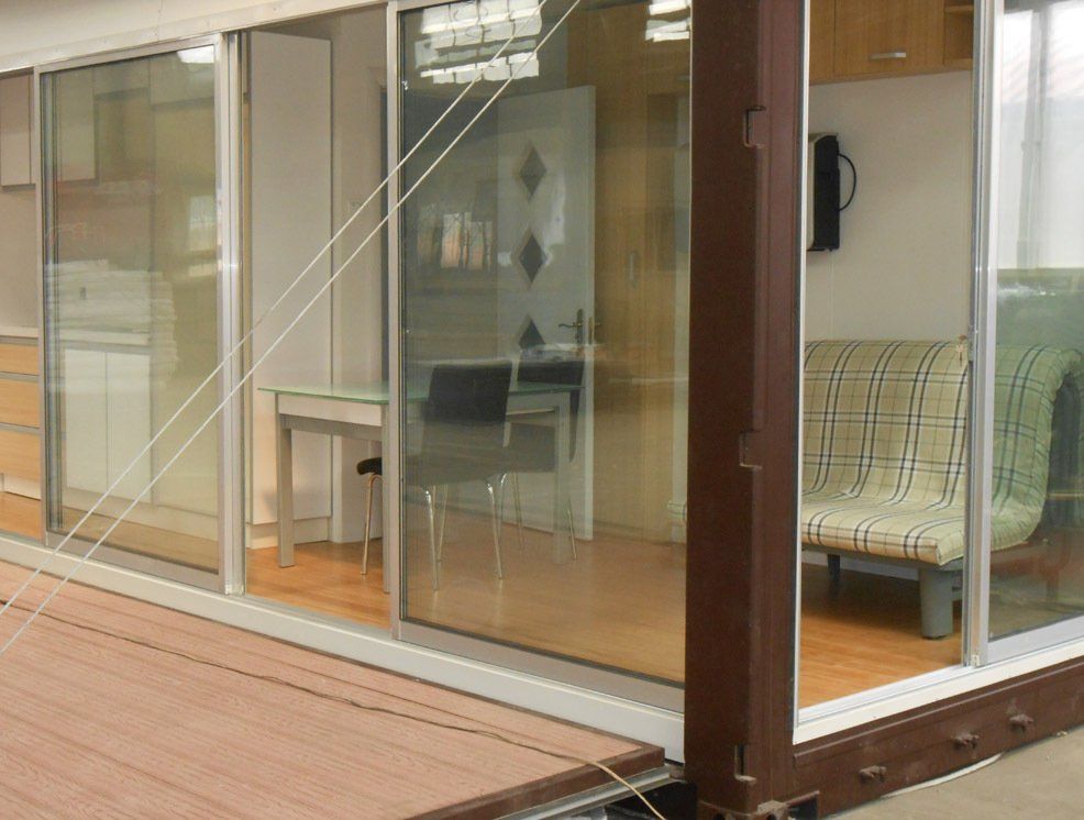 Glass-walled room interior. Brown wooden floor, white walls, desk, sofa visible through glass panels.
