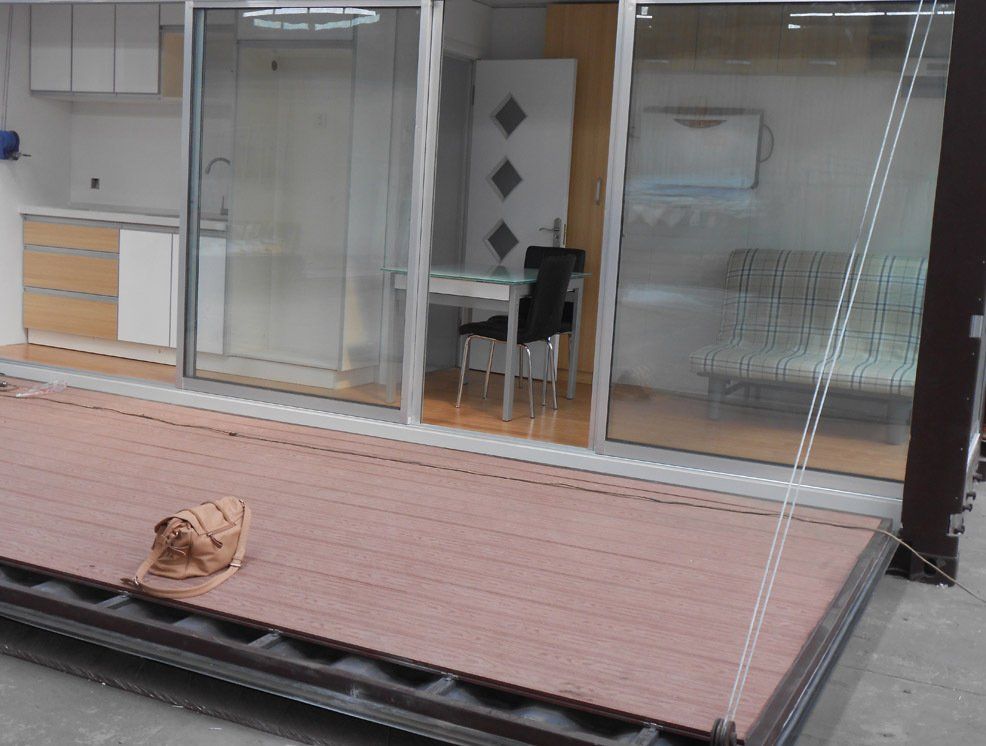 Brown deck leads to a modern container home with sliding doors, showing kitchen, dining table, and bedroom.