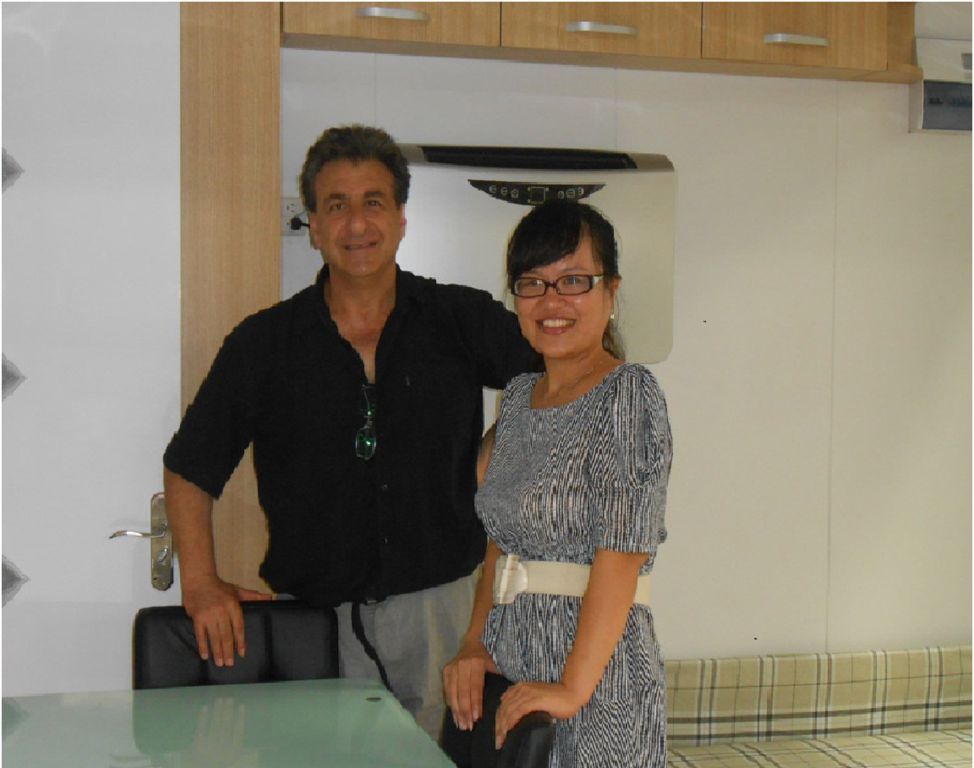 Man and woman smiling, standing in front of a wall with cabinets. The man wears black, the woman a blue patterned dress.