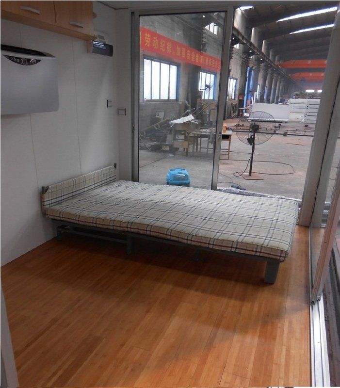 Small room interior with bed, glass door, and wooden floor; factory setting visible outside.