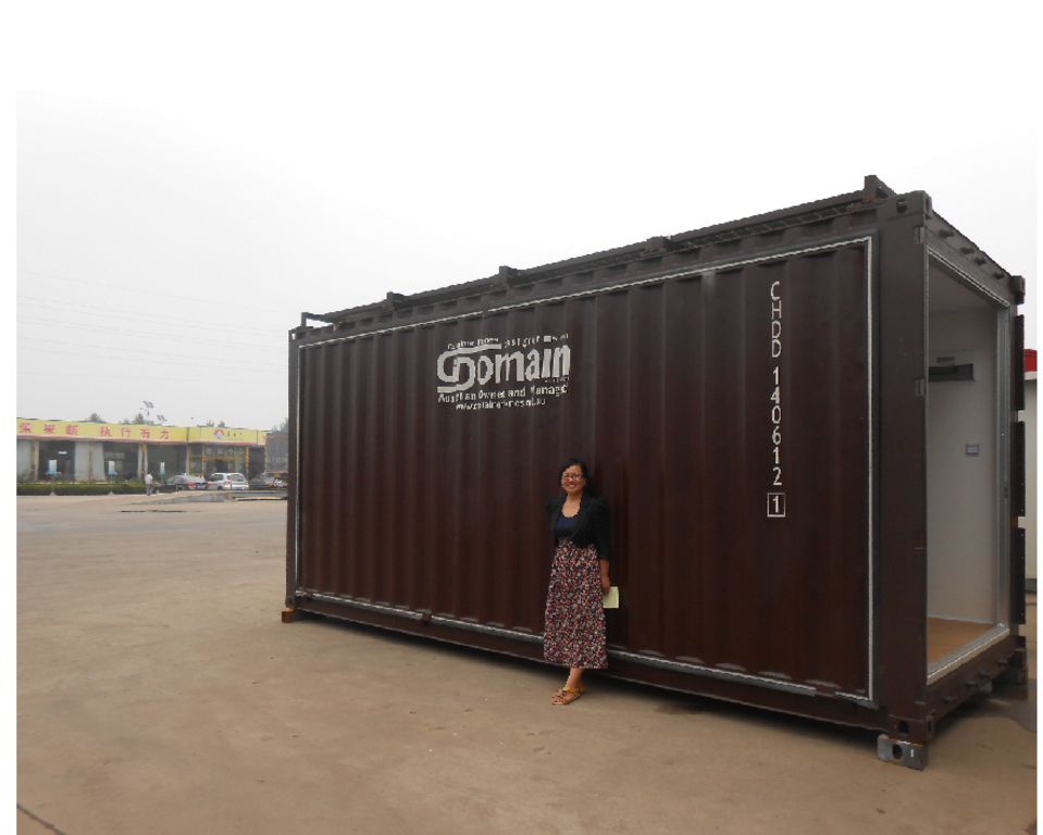 Woman stands beside a large, brown shipping container modified into a building, on a concrete surface.