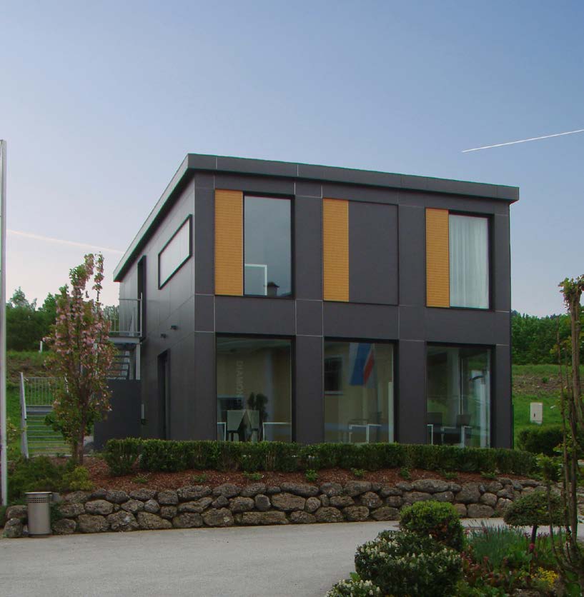 Modern two-story building with dark gray siding, large windows, and wooden accents, set against a green hillside.