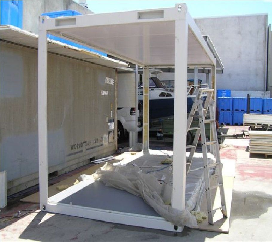 White metal frame being constructed, partially built over a container. Outdoors, daytime.