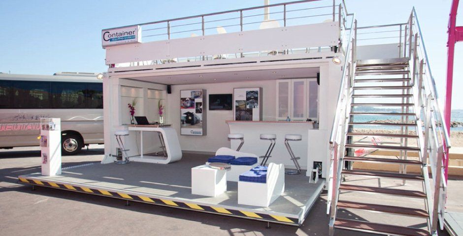 White mobile business unit with rooftop platform, stairs, and seating. Beach setting, bus in background.