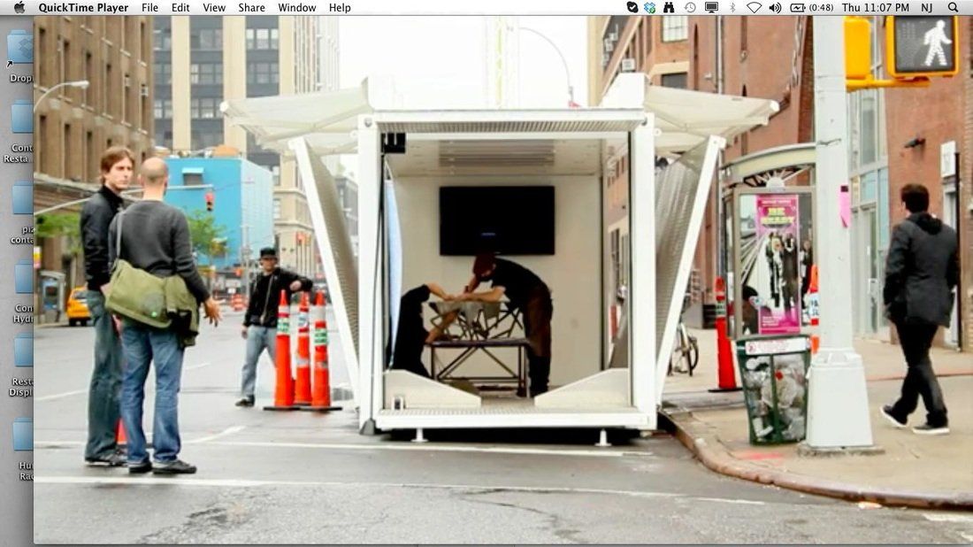 A mobile art installation on a city street features a clear-walled cube with a person inside working on a sculpture.