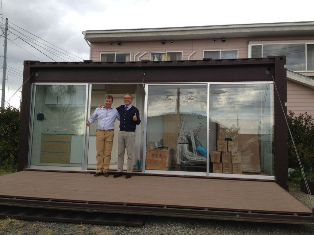 Two men standing in front of a modified shipping container with glass doors. One gives a thumbs-up. Exterior view.