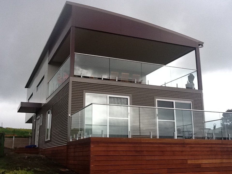 Two-story modern house with brown siding, glass railings, and a wooden deck. Overcast sky.