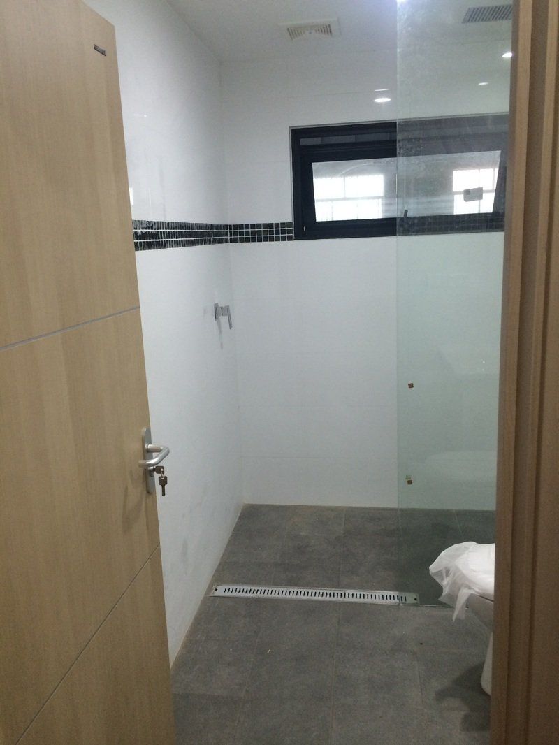 Bathroom interior with light wood door, white walls, gray tile floor, glass shower, and small window.
