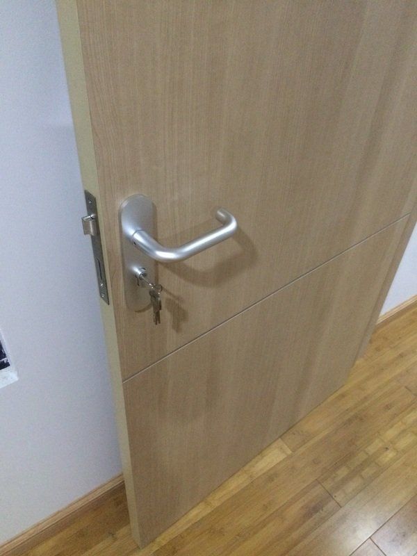 Tan door with silver handle and keys hanging, set against a white wall and wooden floor.