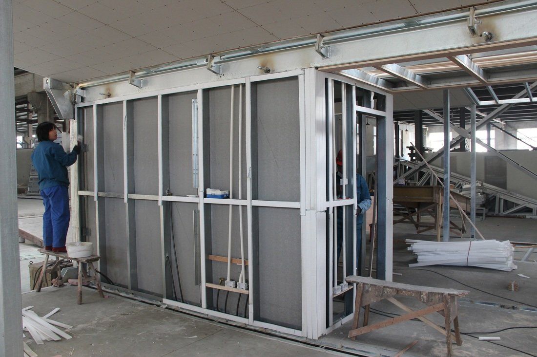 Construction worker installing panels on a metal framed interior wall in a large industrial setting.
