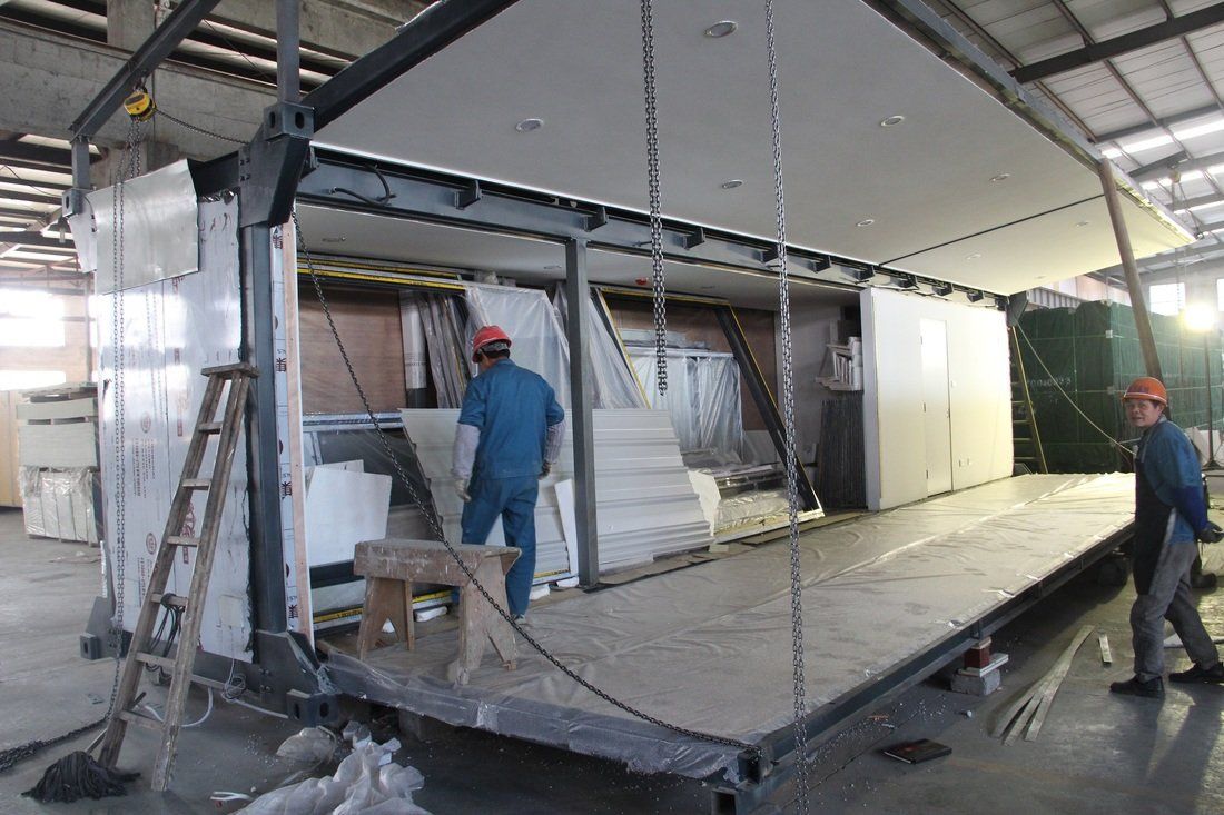 Workers constructing a modular building inside a factory. One worker stands near a small step stool. Another walks toward the edge.